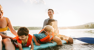 Familia disfrutando de un día soleado en el lago, con niños pequeños usando chalecos salvavidas de color naranja mientras juegan sobre una tabla de paddle surf, acompañados de adultos sonrientes al aire libre.